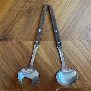 May include: Two vintage serving spoons with wooden handles and silver-colored bowls. One spoon has a rounded bowl, and the other has a unique, open-mouthed design. The handles are dark brown with two small, round, light-colored accents.