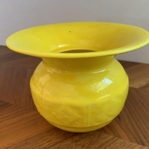 May include: A bright yellow ceramic vase with a wide, flared rim and a rounded body. The vase has a glossy finish and a textured surface, adding visual interest to the design. This decorative piece is suitable for displaying flowers or as a standalone accent.