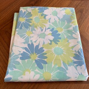 Puede incluir: Tela doblada con un estampado floral de margaritas en blanco, azul claro y verde lima. La tela está doblada y parece ser una sábana u otro artículo de cama. Las margaritas tienen pétalos blancos, azules claros y centros verde lima.