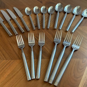 Puede incluir: Un juego de cubiertos de acero inoxidable que incluye cuatro cuchillos, cinco cucharas y seis tenedores. Cada pieza presenta un acabado pulido y liso con un diseño de mango cilíndrico. Los utensilios están dispuestos en filas, mostrando un color plateado metálico uniforme.