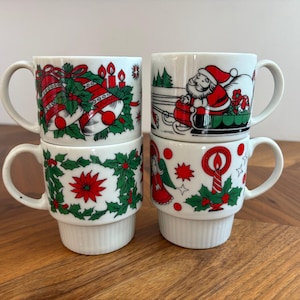May include: Four white ceramic mugs stacked in two columns, each featuring festive Christmas illustrations. Designs include red bells with green holly, Santa Claus in a sleigh, a poinsettia wreath, and an angel with a red candle and holly.