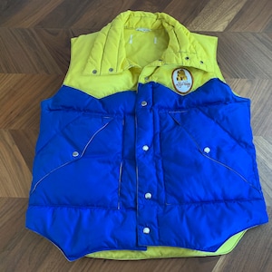 May include: A vintage, quilted vest in bright yellow and blue. The vest has a collared neckline and a patch with text and a logo. It features a diamond-stitched pattern and snap closures. A retro outerwear piece.
