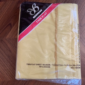 Puede incluir: Una sábana plana amarilla para cama individual en un paquete de plástico. El paquete tiene una etiqueta negra y roja con el texto "Twin Flat" y "No-Iron 50% Fortrel Polyester, 50% Cotton (Cool)". La sábana mide 167 x 243 cm y se adapta a un colchón de 99 x 190 cm.