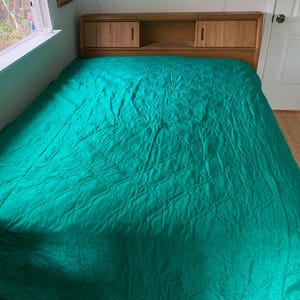 Op de afbeelding: Een bed met een turquoise groene sprei. Het bed heeft een houten hoofdbord met twee kleine kastdeuren en een plank. Het bed staat in een kamer met een raam en een deur.
