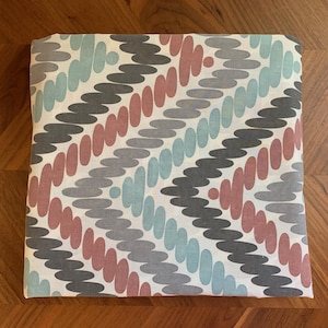 May include: Folded fabric with a repeating pattern of abstract shapes in shades of gray, dusty rose, and teal on a white background. The fabric is rectangular and appears to be made of cotton or a similar material.