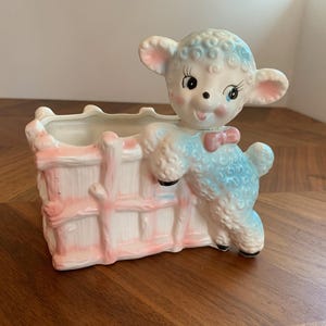 Può includere: Un vaso di ceramica vintage con un agnello appoggiato a una staccionata rosa e bianca. L'agnello ha accenti blu, un papillon rosa e un'espressione dolce. Questo vaso decorativo è ideale per piccole piante o fiori.