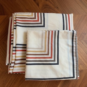 May include: A folded fabric textile featuring a geometric pattern of concentric right-angled lines in shades of cream, rust orange, mustard yellow, dark brown, and black. The design consists of repeating linear borders that create a structured, layered appearance across the surface of the material.