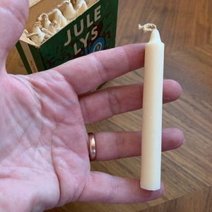 Pu&ograve; includere: Una candela color avorio con una miccia attorcigliata &egrave; tenuta in mano. La candela &egrave; cilindrica. Una scatola verde con la scritta "JULE LYS" &egrave; parzialmente visibile sullo sfondo, suggerendo un tema festivo. La candela &egrave; lunga circa 10 cm.