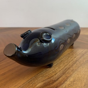 May include: A navy blue ceramic piggy bank with a glossy finish. The bank has a slot on top for coins and small, circular accents along its body. It has a rounded snout, small ears, and three small feet.
