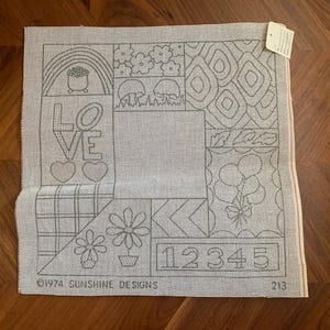 Könnte beinhalten: Eine quadratische, cremefarbene Gobelin-Leinwand mit verschiedenen umrissenen Designs, darunter ein Regenbogen, Blumen, Pilze, Luftballons und das Wort "LOVE". Die Zahlen "12345" sind ebenfalls vorhanden. Der Text "©1974 SUNSHINE DESIGNS" ist unten aufgedruckt.