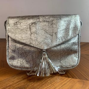 May include: Silver-colored crossbody bag with a flap closure and a decorative tassel. The bag has a textured, crackled appearance. The bag is sitting on a wooden surface.