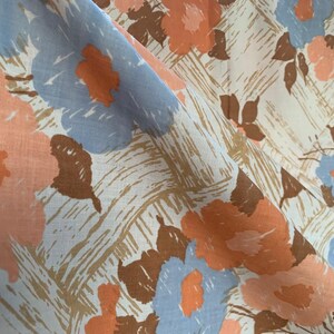 May include: Close-up of a fabric with a floral pattern. The design features large flowers in shades of peach, light blue, and brown against a cream-colored background with a textured, cross-hatched pattern. The fabric is folded, showing its texture.