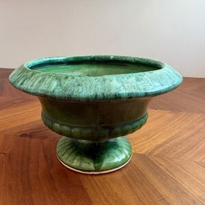 May include: A ceramic pedestal bowl featuring a deep green glaze with lighter teal and emerald streaks. The bowl has a wide, flared rim, a rounded body with a subtle horizontal ribbed texture, and a circular base with radiating fluted details.