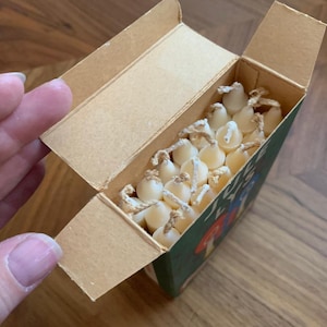Pu&ograve; includere: Una scatola di cartone parzialmente aperta, piena di piccole candele color crema. La scatola ha un lato verde con un'illustrazione stravagante. Le candele hanno uno stoppino di corda. Una mano tiene aperta la scatola.