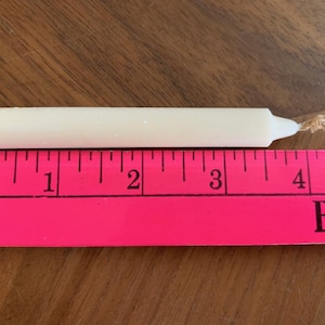 Pu&ograve; includere: Una candela affusolata color crema, lunga circa 10 cm, &egrave; appoggiata su una superficie di legno accanto a un righello rosa. La candela ha uno stoppino a un'estremit&agrave;. Il righello &egrave; contrassegnato da linee e numeri neri.