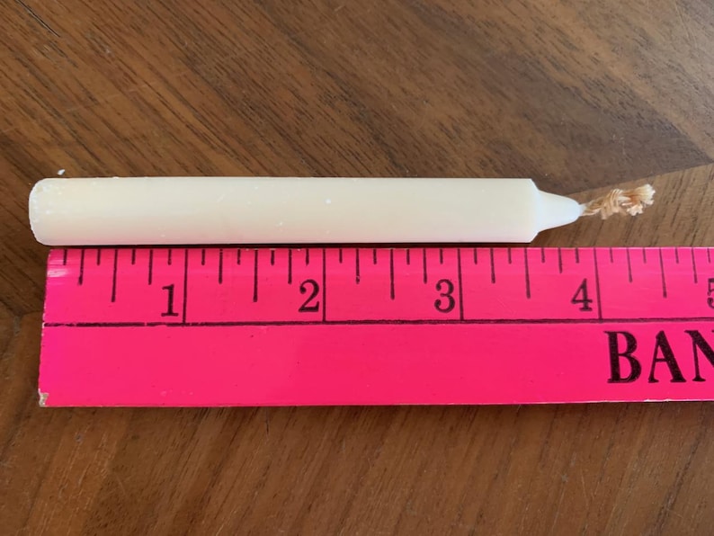Pu&ograve; includere: Una candela affusolata color crema, lunga circa 10 cm, &egrave; appoggiata su una superficie di legno accanto a un righello rosa. La candela ha uno stoppino a un'estremit&agrave;. Il righello &egrave; contrassegnato da linee e numeri neri.