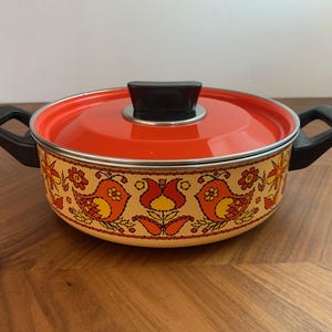Puede incluir: Una olla de cocina vintage naranja y crema con tapa. La olla presenta un patrón decorativo de pájaros y flores en naranja, amarillo y rojo. Tiene asas negras y un pomo negro en la tapa.