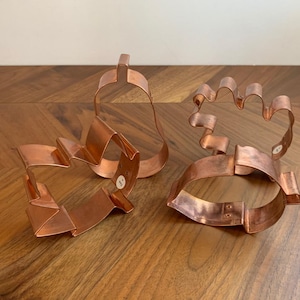 May include: Three copper cookie cutters in the shapes of a heart, a pear, and a hand. The heart and hand cutters have folded edges. The pear cutter has a loop at the top. All three are on a wooden surface.