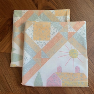 May include: Two folded bed sheets with a pastel patchwork design. The pattern includes geometric shapes, a sun, and houses in shades of pink, yellow, and blue. The sheets are on a wooden surface.