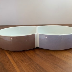 Puede incluir: Un cuenco para servir dividido con un diseño moderno. Un lado es marrón apagado, el otro lavanda suave. El interior es blanco. El cuenco tiene forma ovalada y se apoya sobre una superficie de madera.