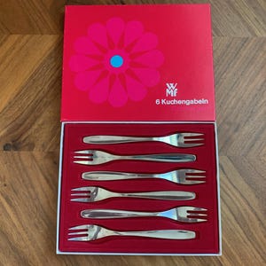 May include: A set of six stainless steel cake forks presented in a red box. The box features a large pink flower design with a blue center and the text "6 Kuchengabeln". The forks are arranged on a red velvet-like interior.