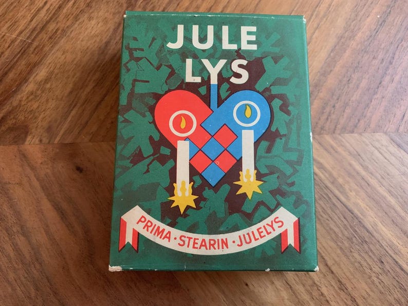 Pu&ograve; includere: Una scatola verde con le parole "JULE LYS" in bianco. La scatola presenta un disegno a forma di cuore in rosso e blu con due candele e il testo "PRIMA STEARIN JULELYS" su uno striscione. Lo sfondo ha un motivo di foglie verdi.