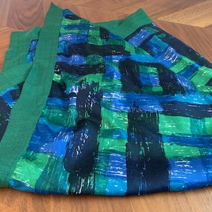 May include: A green and blue patterned fabric, possibly a scarf or shawl, lies on a wooden surface. The fabric features a bold design with brushstroke-like patterns in shades of green, blue, and black. The edges are trimmed with a solid green border.