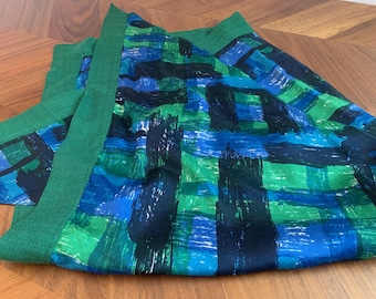 May include: A green and blue patterned fabric, possibly a scarf or shawl, lies on a wooden surface. The fabric features a bold design with brushstroke-like patterns in shades of green, blue, and black. The edges are trimmed with a solid green border.
