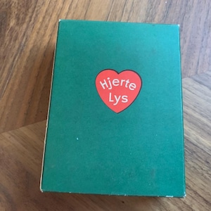 Pu&ograve; includere: Una scatola verde con un cuore rosso con la scritta bianca "Hjerte Lys". La scatola &egrave; su una superficie di legno.
