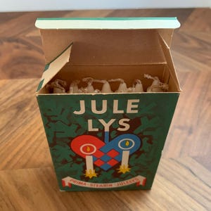 Pu&ograve; includere: Una scatola vintage di candele Jule Lys, con la scritta "JULE LYS" in bianco. La scatola &egrave; verde con un disegno a forma di cuore con due candele. La scatola &egrave; aperta, rivelando le candele all'interno.