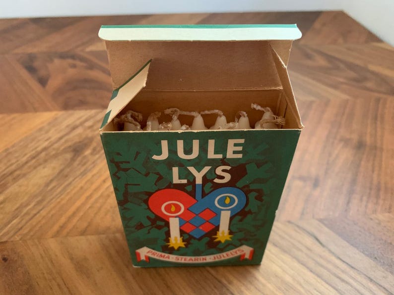 Pu&ograve; includere: Una scatola vintage di candele Jule Lys, con la scritta "JULE LYS" in bianco. La scatola &egrave; verde con un disegno a forma di cuore con due candele. La scatola &egrave; aperta, rivelando le candele all'interno.