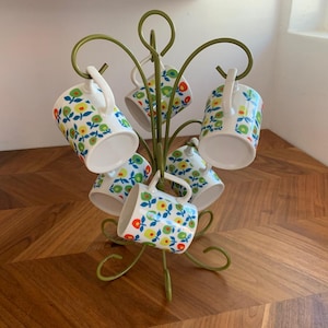 May include: A set of six white ceramic mugs with a floral pattern of blue, yellow, and red flowers and green leaves. The mugs are displayed on a green metal mug tree with a curved design. The mugs are hanging from the tree.