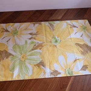 Puede incluir: Una tela floral rectangular con grandes flores amarillas y blancas con acentos verdes y marrones. Las flores están pintadas sobre un fondo amarillo. La tela es probablemente un mantel o un artículo de decoración del hogar similar.