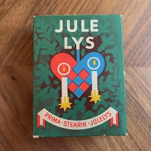 Pu&ograve; includere: Scatola rettangolare verde con le parole "JULE LYS" in bianco. La scatola presenta un disegno a forma di cuore rosso e blu con due candele e il testo "PRIMA - STEARIN - JULELYS" su uno striscione.