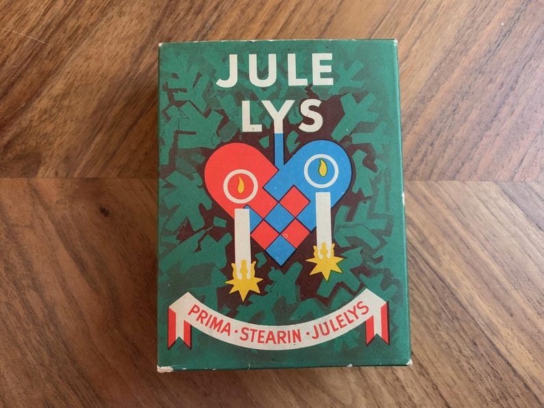 Pu&ograve; includere: Scatola rettangolare verde con le parole "JULE LYS" in bianco. La scatola presenta un disegno a forma di cuore rosso e blu con due candele e il testo "PRIMA - STEARIN - JULELYS" su uno striscione.