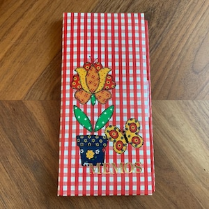 May include: A red and white gingham-patterned memo pad with a floral and butterfly design. The cover features a flower in a blue pot, a butterfly, and the word "MEMOS" in gold lettering. The memo pad is closed and rectangular.