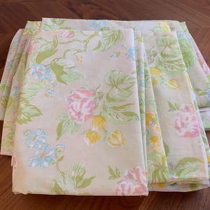 Pode incluir: Um conjunto de lençóis de cama dobrados com um padrão floral. Os lençóis apresentam um fundo creme adornado com um padrão repetido de flores e folhas rosa, amarelas, azuis e verdes. Os lençóis estão cuidadosamente dobrados e empilhados.