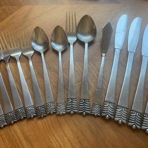 Puede incluir: Un conjunto de cubiertos antiguos, que incluye tenedores, cucharas, cuchillos de mantequilla y cuchillos de mesa. La cubertería presenta un diseño decorativo en los mangos, dispuestos sobre una superficie de madera.