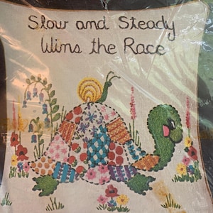 May include: A square fabric wall hanging featuring an embroidered turtle with a colorful patchwork shell, a small snail on its back, and surrounding flowers. The text "Slow and Steady Wins the Race" is embroidered in dark brown thread at the top.