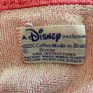 Może przedstawiać: Zbliżenie na r&oacute;żowo-białą tkaninę o fakturze z etykietą z napisem "A Disney exclusive 100% Cotton-Made in Brazil". Etykieta zawiera r&oacute;wnież instrukcje dotyczące pielęgnacji.