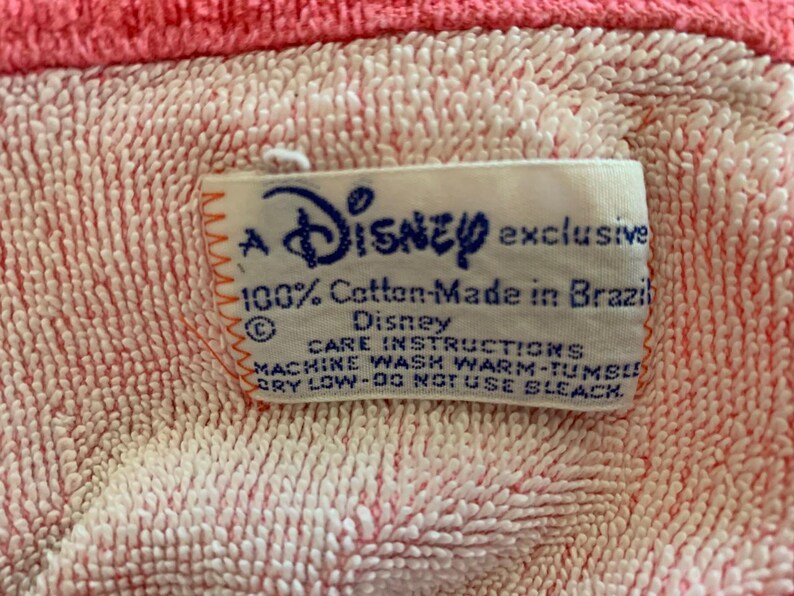 Może przedstawiać: Zbliżenie na r&oacute;żowo-białą tkaninę o fakturze z etykietą z napisem "A Disney exclusive 100% Cotton-Made in Brazil". Etykieta zawiera r&oacute;wnież instrukcje dotyczące pielęgnacji.