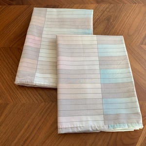 Peut inclure: Deux draps de lit pliés avec un motif de blocs de couleurs pastel. Les draps présentent des rayures horizontales dans les tons de rose, de bleu et de gris. Les draps sont en tissu doux et léger, pliés sur une surface en bois.