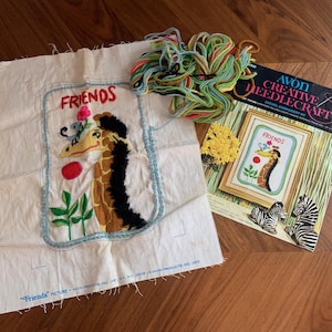 May include: An embroidery kit featuring a giraffe design with the word "FRIENDS". The kit includes a canvas with the giraffe design, colorful yarn, and an "Avon Creative Needlecraft" booklet. The image also shows a small yellow flower and zebra figurines.