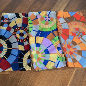 May include: Three rectangular silk scarves featuring a repeating geometric pattern of overlapping circles. The first scarf displays black, blue, red, and yellow tones. The second scarf shows green, purple, and yellow. The third scarf features orange, brown, and blue.