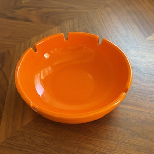 Könnte beinhalten: Ein leuchtend orangefarbener, kreisförmiger Aschenbecher mit glatter, glänzender Oberfläche. Die Schale verfügt über drei gleichmäßig verteilte Einkerbungen am Rand, die zum sicheren Ablegen dienen. Das Objekt hat eine einfache, runde Form.