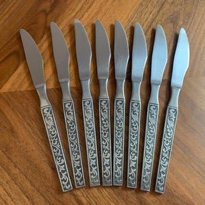 Puede incluir: Juego de ocho cuchillos de mesa de acero inoxidable. Cada cuchillo presenta una hoja plateada y un mango con un diseño floral grabado. Los cuchillos están dispuestos sobre una superficie de madera, mostrando su elegante diseño.