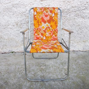 Peut inclure: Chaise pliante vintage avec une structure métallique et des accoudoirs en bois. L'assise et le dossier présentent un motif floral vibrant dans les tons orange, jaune et rose. La chaise est conçue pour une utilisation en extérieur et a une esthétique rétro classique.
