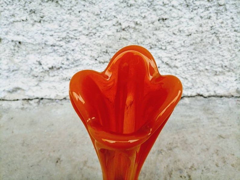 May include: A vibrant orange glass vase with a unique, organic shape. The vase has a flared top with rounded edges, and a smooth, glossy finish. The interior is a deep orange, creating a striking contrast.