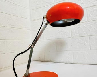 Könnte beinhalten: Eine rote Metall-Tischlampe mit einem runden Schirm und einem verstellbaren Arm aus Silber. Die Lampe hat ein schwarzes Kabel und einen runden Sockel.