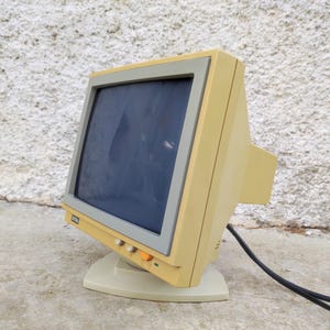 Op de afbeelding: Een vintage beige beeldbuiscomputermonitor met een grijze schermrand. De monitor heeft een vierkant display, een stevige voet en een opvallende oranje aan-uitknop op het voorpaneel, met een zwarte stroomkabel die aan de achterkant uitsteekt.
